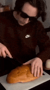 a man wearing sunglasses is cutting a loaf of bread with a knife on a cutting board .